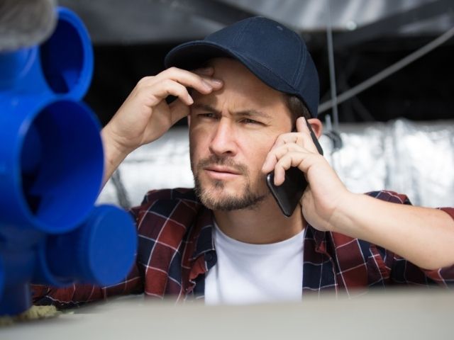 Person holding a mobile phone beside stacked blue pipes at a construction site, illustrating trade support, technical assistance and professional advice for building and installation projects.