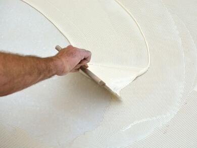 Hand using a notched trowel to spread Evostik flooring adhesive across a subfloor, creating a consistent ribbed pattern ready for the installation of floor coverings.