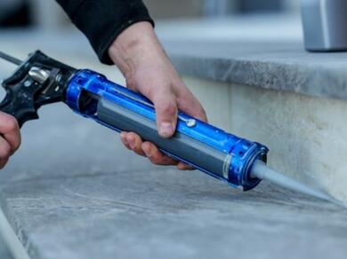 Person applying sealant with a cartridge gun along the joint between a stone-effect worktop and upstand, illustrating how to choose the right sealant for durable kitchen and interior sealing applications.