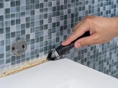 Hand using a scraper tool to remove old silicone sealant from the joint between a tiled wall and a worktop, preparing the surface for a new sealant application.