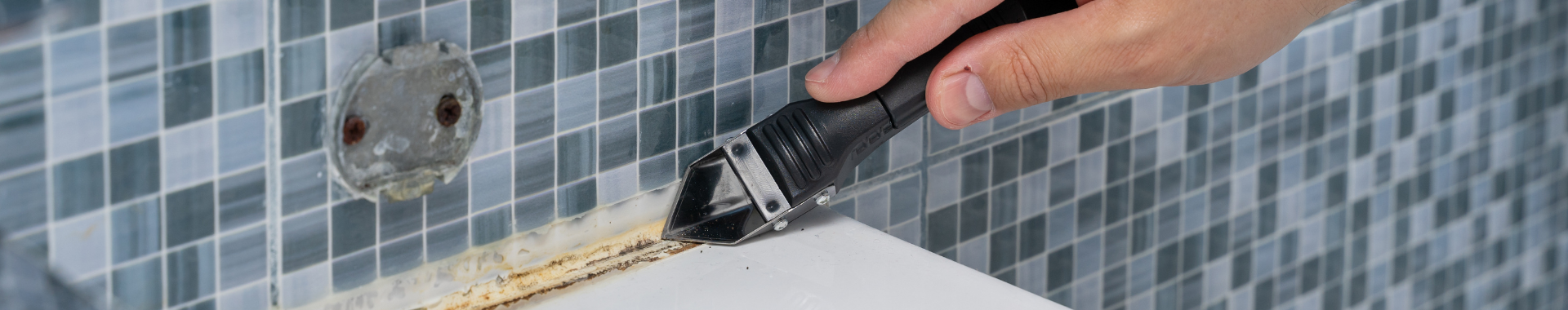 A man uses a scraper to remove old, discolored silicone sealant from the edge of a bathtub
