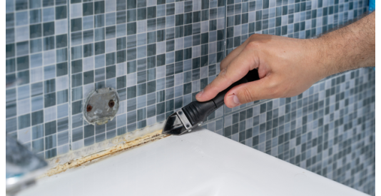 A man uses a scraper to remove old, discolored silicone sealant from the edge of a bathtub