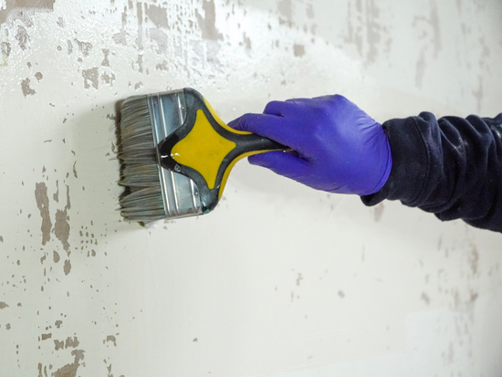 A hand in a blue glove uses a wide brush to apply Contractors PVA on a speckled, textured wall